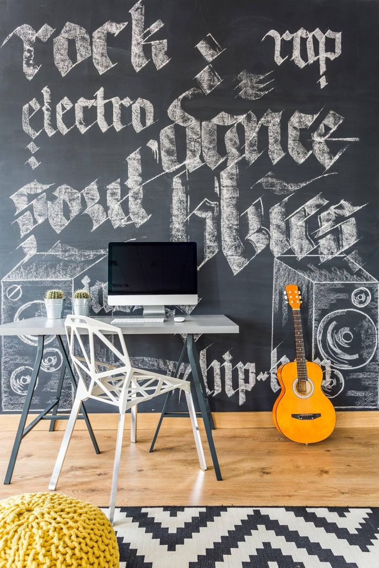 a desk with a computer and a guitar in front of a chalkboard wall